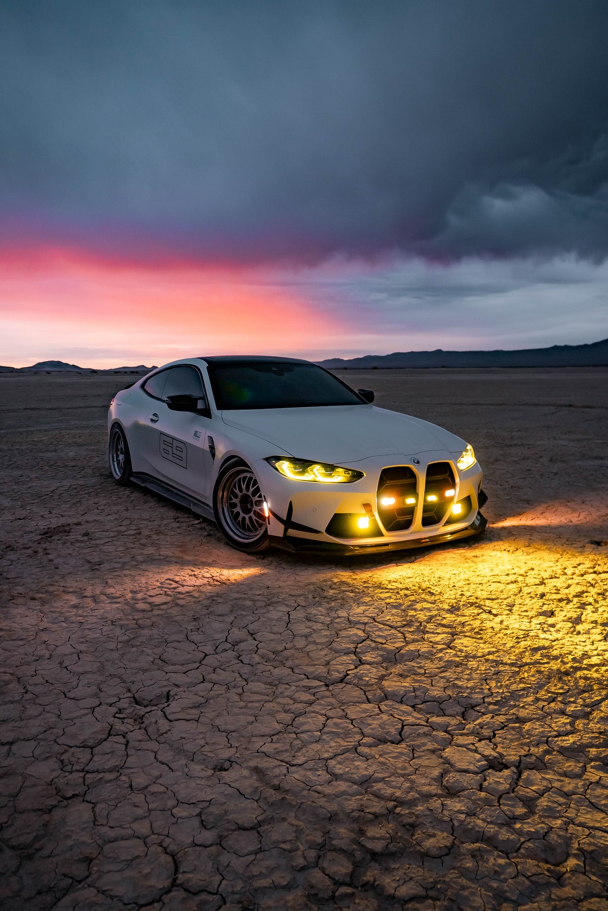 White coupe with amber auxiliary lights illuminated on a dry lakebed at sunset